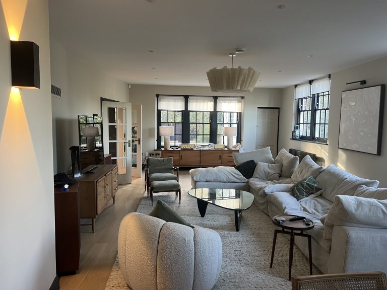 Lewis Capaldy – A spacious living room with a large cream sectional sofa mid-century wooden furniture black-framed windows a sculptural ceiling light and a glass coffee table on a textured rug