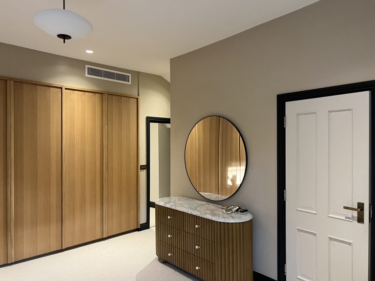 Lewis Capaldy-A modern hallway or dressing area with built-in wooden wardrobes a round wall mirror above a marble-topped sideboard beige walls and a white panel door with a brass handle