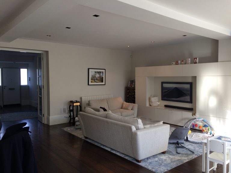 Sunette –A modern living room with a light grey sofa built-in wall cabinets a mounted TV dark wooden floors and baby play items in the corner